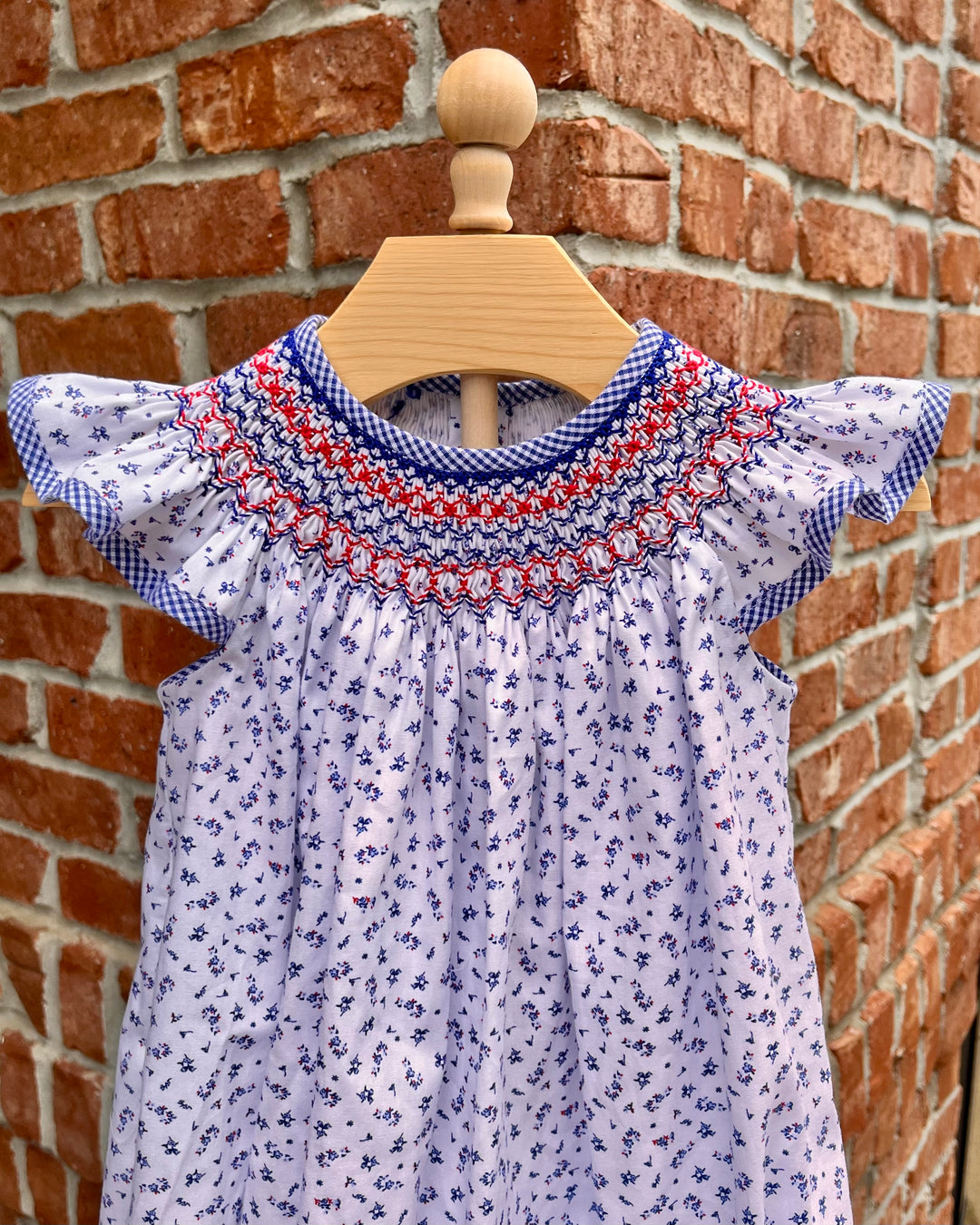 White dress with blue floral pattern and red accents on a wooden hanger against a brick wall.