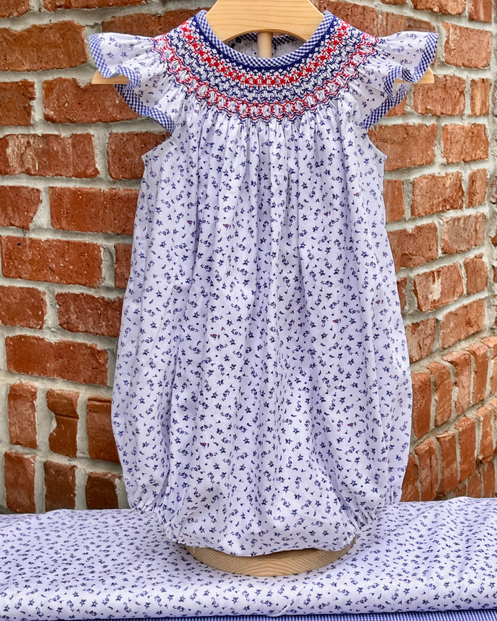 Baby dress with floral pattern and red, white, and blue trim on a wooden hanger against a brick wall.