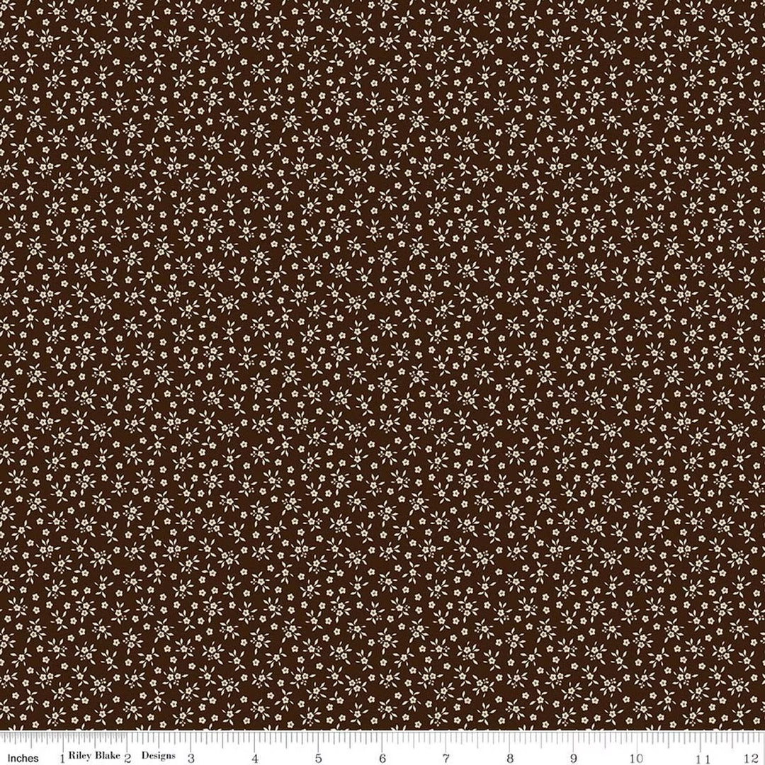 A close-up of a repeating fabric pattern featuring small white floral and leaf motifs on a dark brown background, with a ruler strip along the bottom edge showing scale.