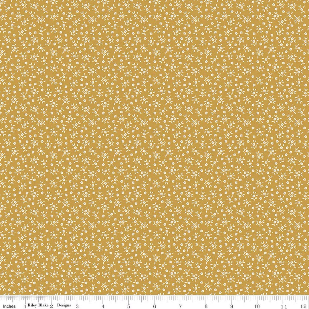 A close-up of a fabric pattern showing a mustard yellow background with a dense, small-scale white floral and leaf print. A white ruler at the bottom edge displays the brand 'Riley Blake Designs' and inch measurements.