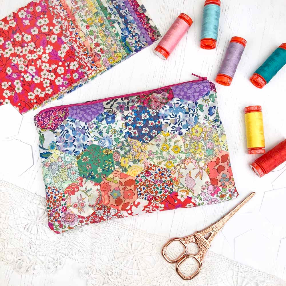 A colorful patchwork fabric zip pouch with a floral and geometric print, styled on a white lace surface with sewing thread spools and scissors.
