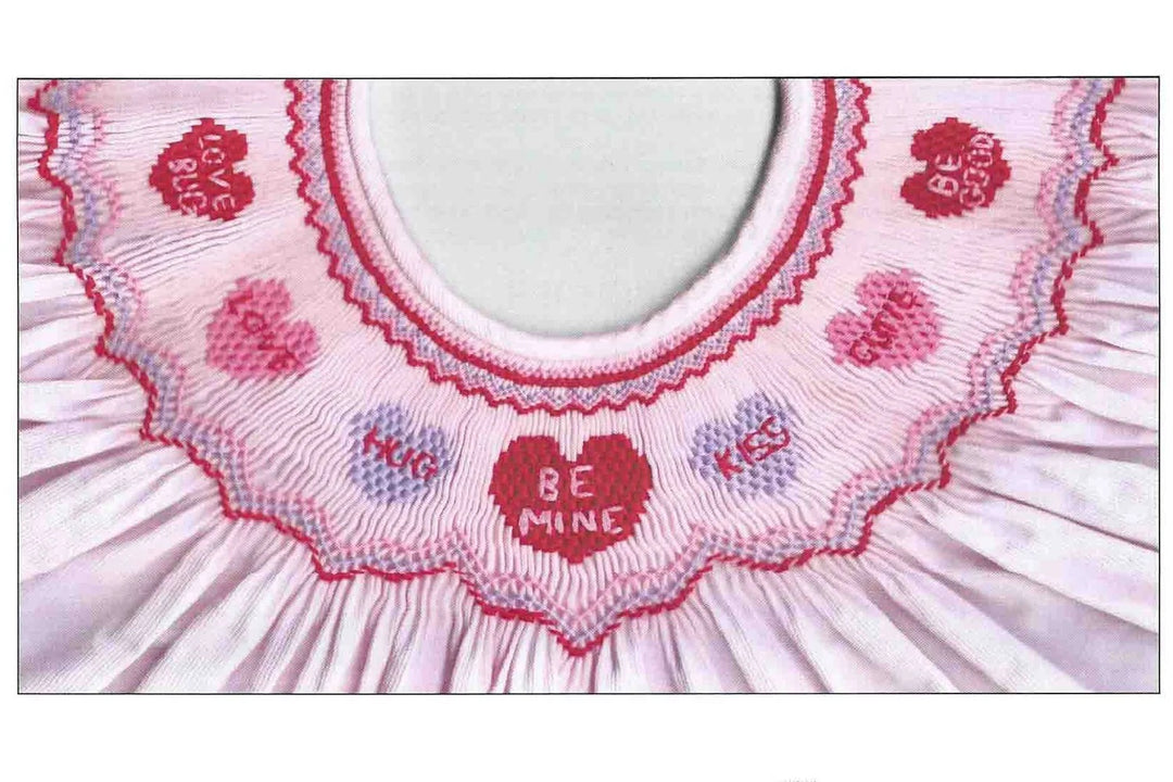 A close-up of a light pink pleated fabric collar with embroidered hearts and the words 'LOVE BE', 'BE ME', and 'KIS' in red and lavender thread.