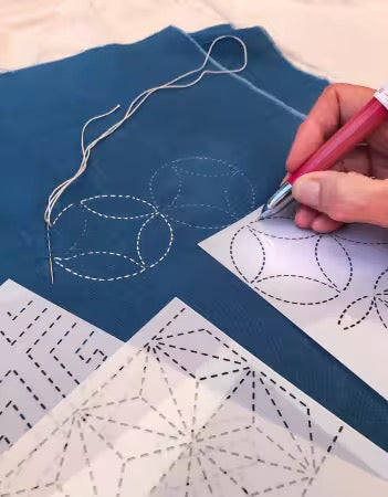 A close-up of a hand using a pink pen to trace geometric quilt patterns onto white paper, with blue fabric and a needle threaded nearby.