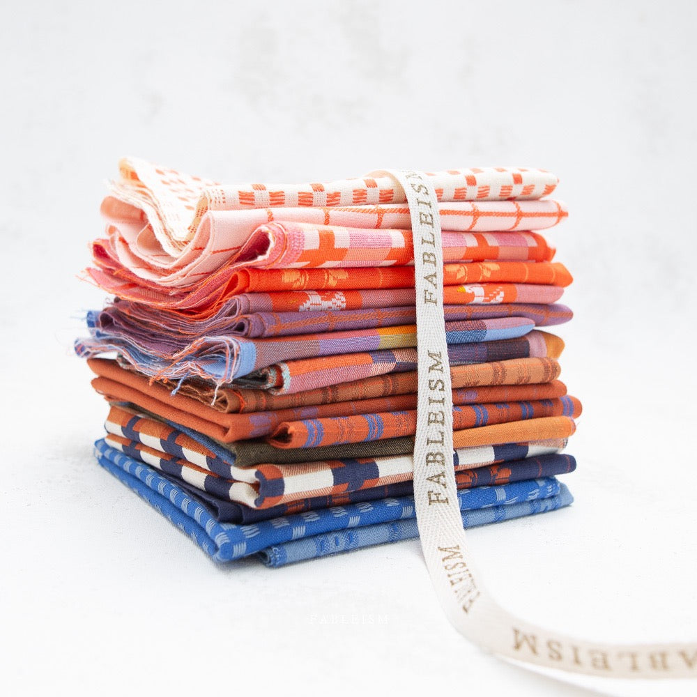 A stack of folded patterned fabric pieces in orange, pink, purple, blue, and brown tones, tied with a white ribbon printed with the word 'FABLEISM'.