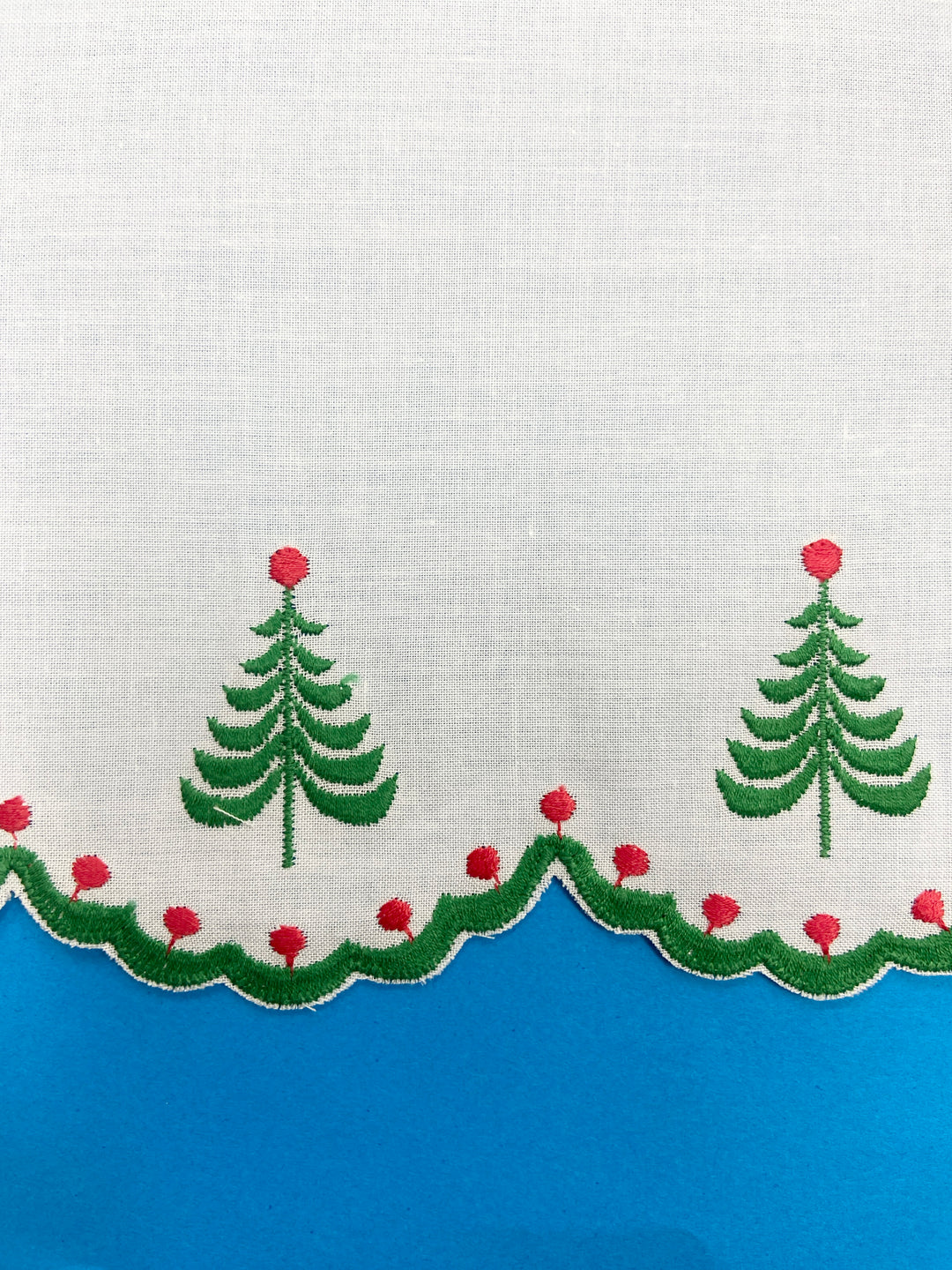 A close-up of a white embroidered sows trim with embroidered Christmas tree and garland motifs along the scalloped edge, set against a bright blue background.