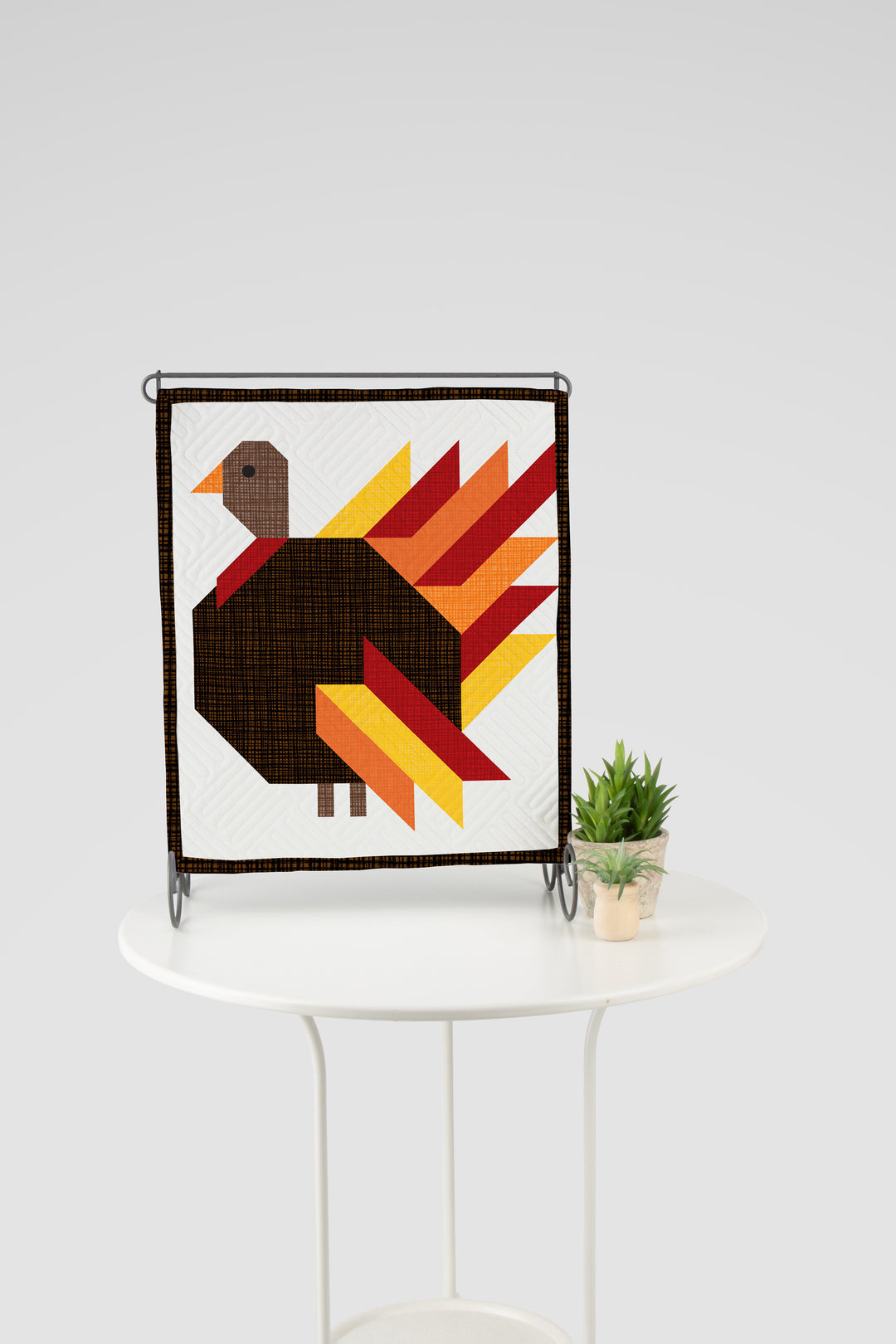 Framed quilt with a colorful turkey design on a white table.
