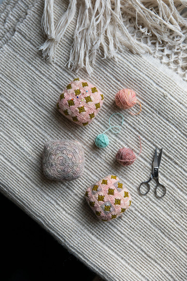 Sitting on a white ribbed table cloth are 3 small crocheted cushions in a variety of pinks and greens and purples.