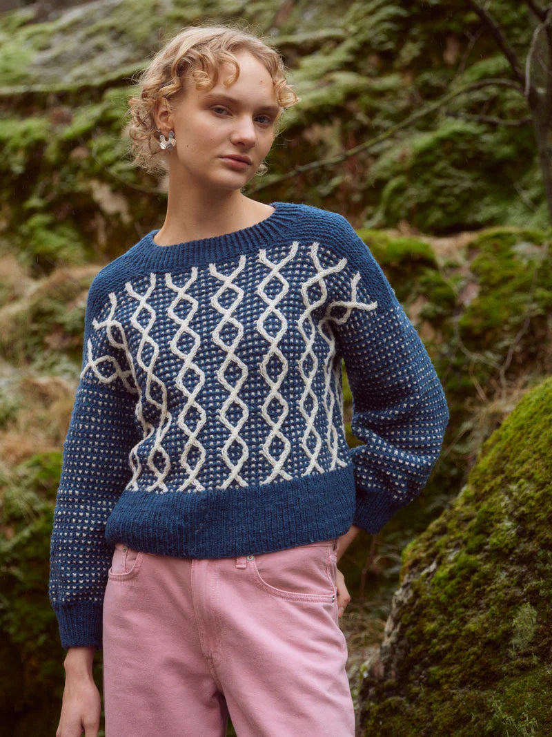 A young white woman with short blonde curly hair stands in front of moss covered rocks. She's wearing a navy blue and white cabled sweater and pink pants.