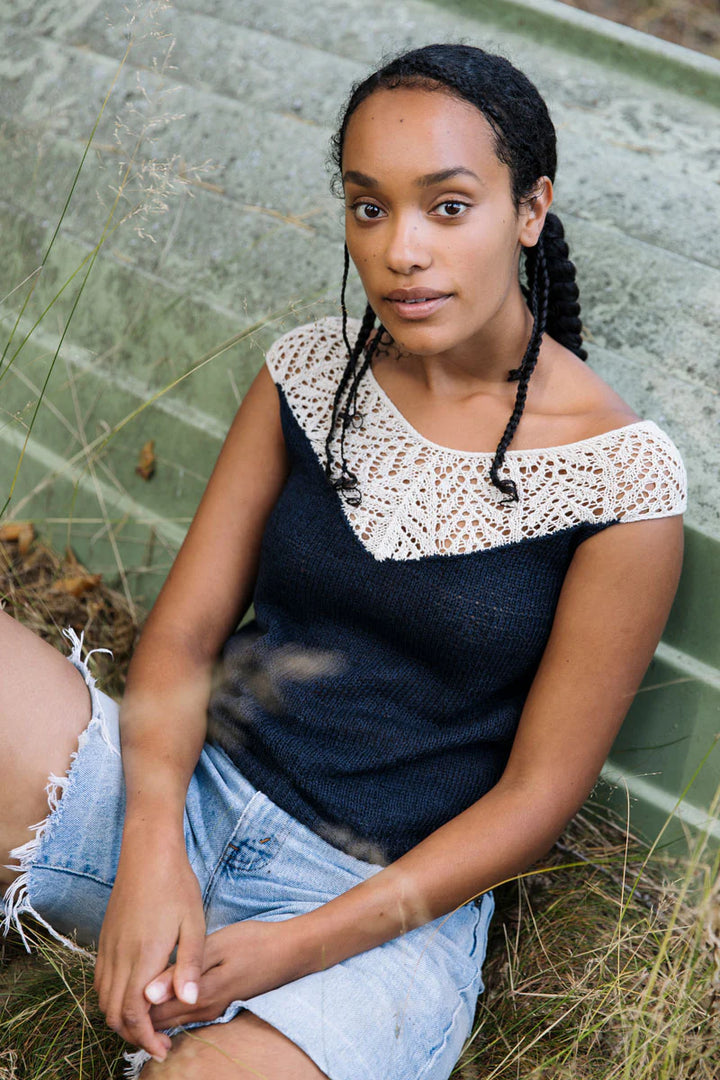 a young black woman wears a black knitted top lined with white lace on the top, sitting in the grass leaning against an overturned boat