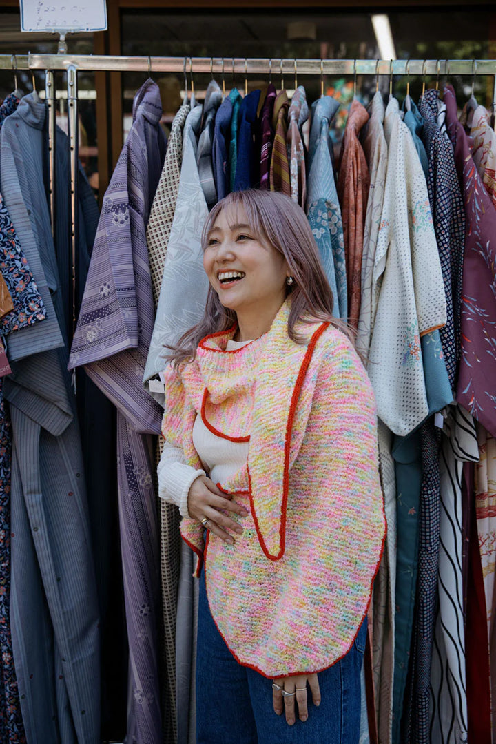 A young Japanese woman wears a multicolored neon shawl with a red edging. She's smiling and looking into the distance and standing in front of a rack of clothing.