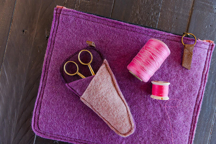 Purple felt pouch with sewing tools including scissors and spools of thread on a wooden surface.
