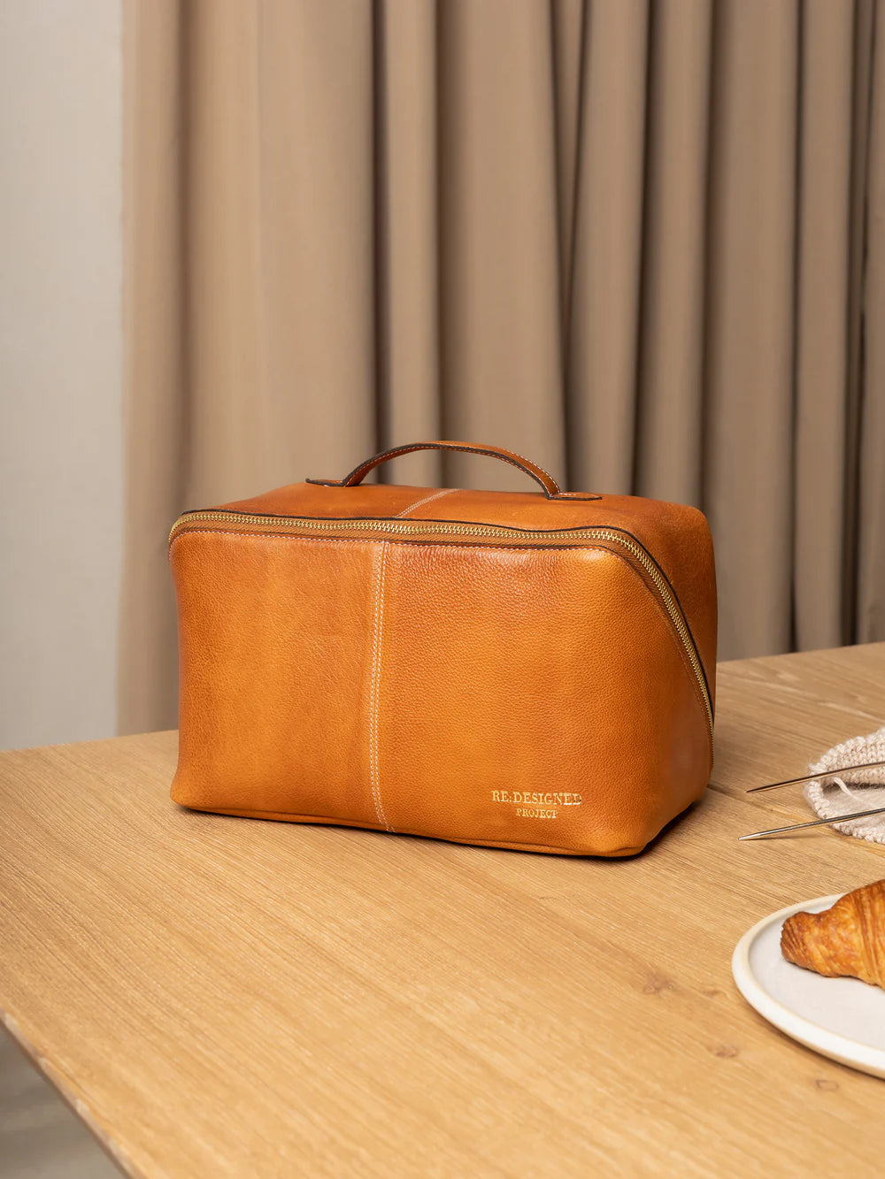 Brown leather toiletry bag on a wooden surface with a plate of food and a curtain in the background.