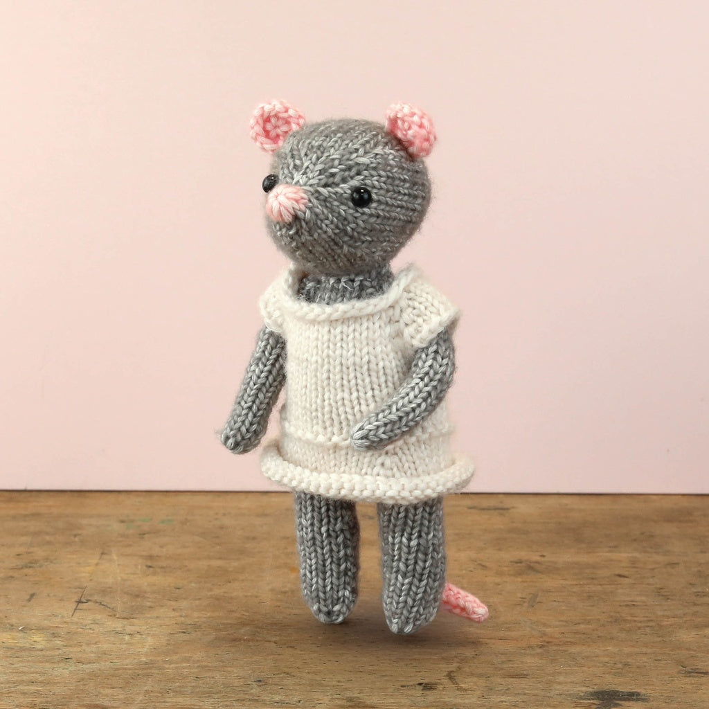 Knitted mouse toy wearing a white dress on a wooden surface with a pink background
