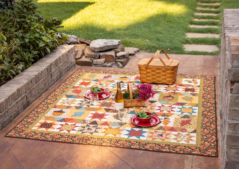 Outdoor setting with a colorful quilt, picnic basket, and tableware on a patio.