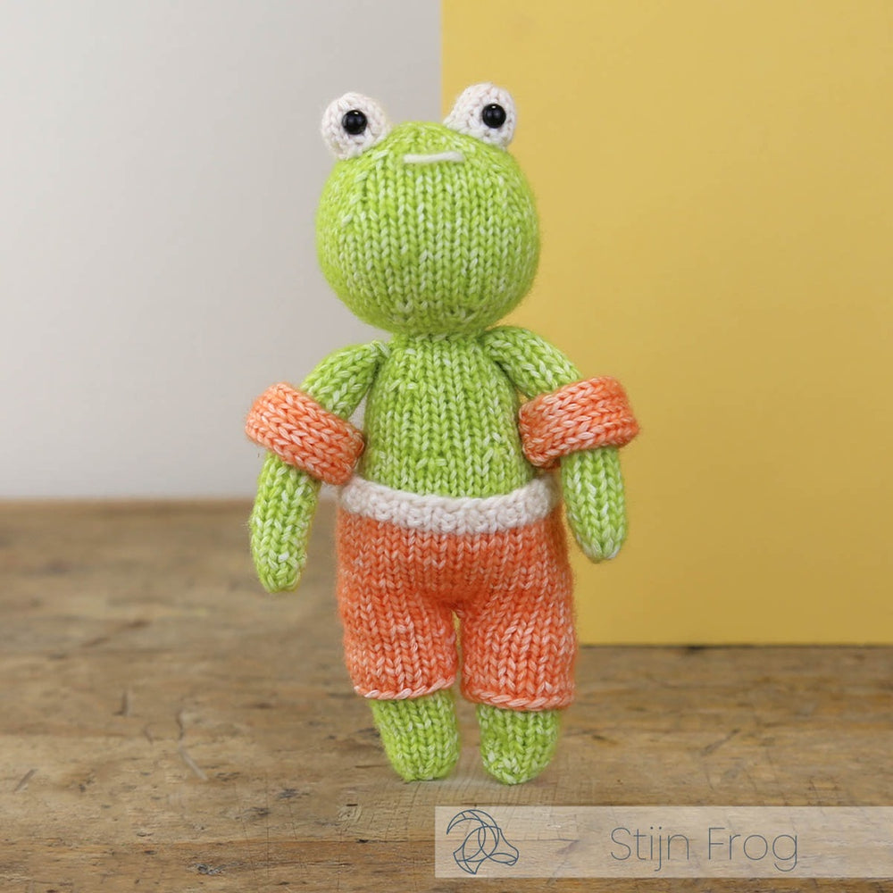 Knitted frog toy with orange shorts on a wooden surface with a yellow wall background