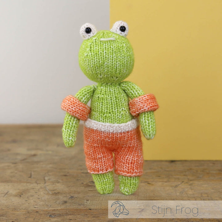 Knitted frog toy with orange shorts on a wooden surface with a yellow wall background