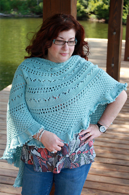 Woman wearing a light blue crocheted poncho by a body of water.