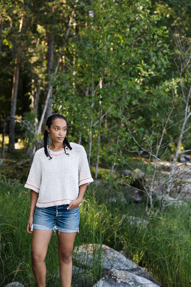 a young black woman wearing a short sleeve white knitted top stands in the woods