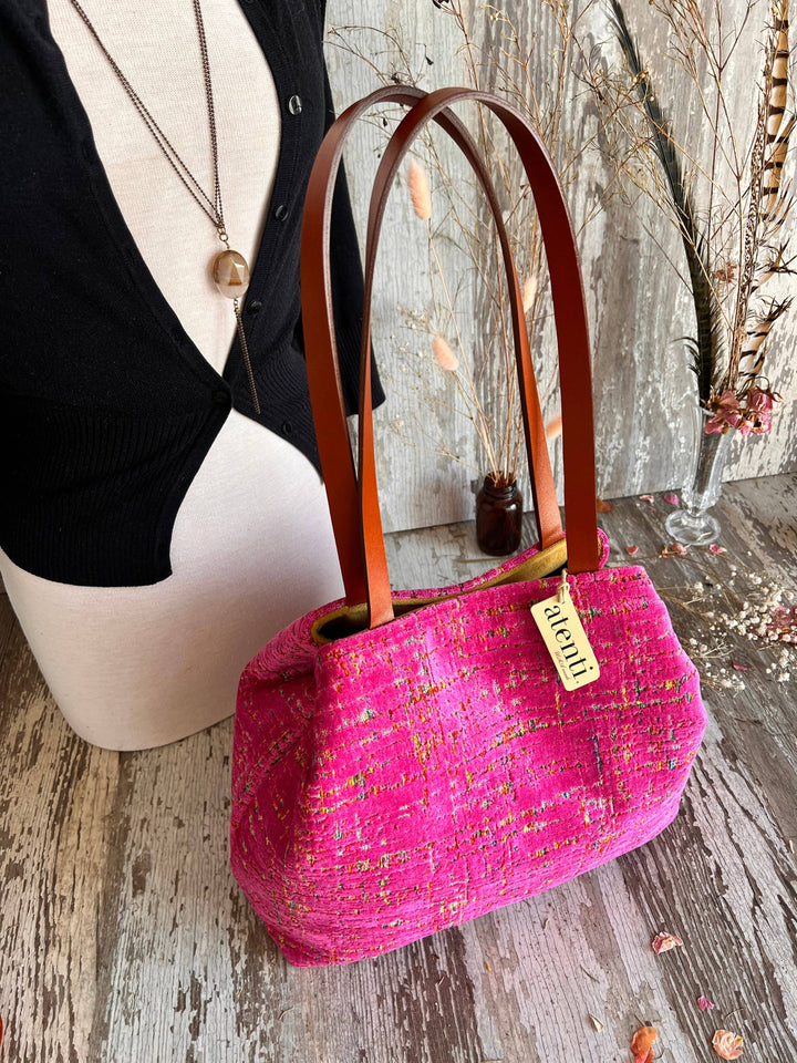 A bright pink textured tweed handbag with brown leather shoulder straps, displayed on a rustic wooden surface next to a mannequin torso.