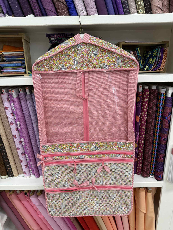 Pink garment bag with floral trim hanging on a rack with fabric in the background