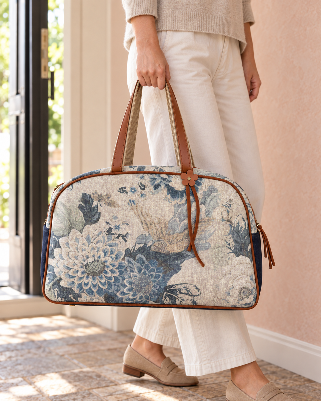 A person in a beige sweater and white pants holds a large duffel bag with a blue and white floral print, brown leather handles, and brown trim.
