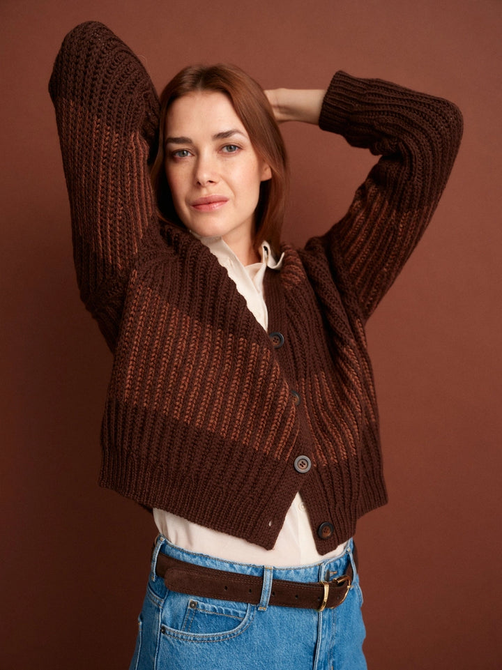 Woman wearing a brown knitted cardigan over a white shirt with blue jeans on a brown background