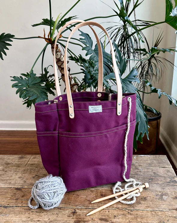 Purple tote bag with beige handles on a wooden surface with yarn and knitting needles, surrounded by plants.
