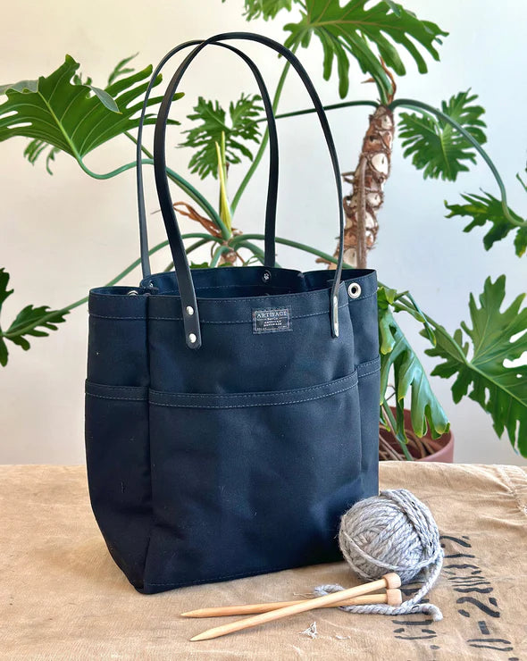 Black tote bag with knitting needles and yarn on a wooden surface with plants in the background