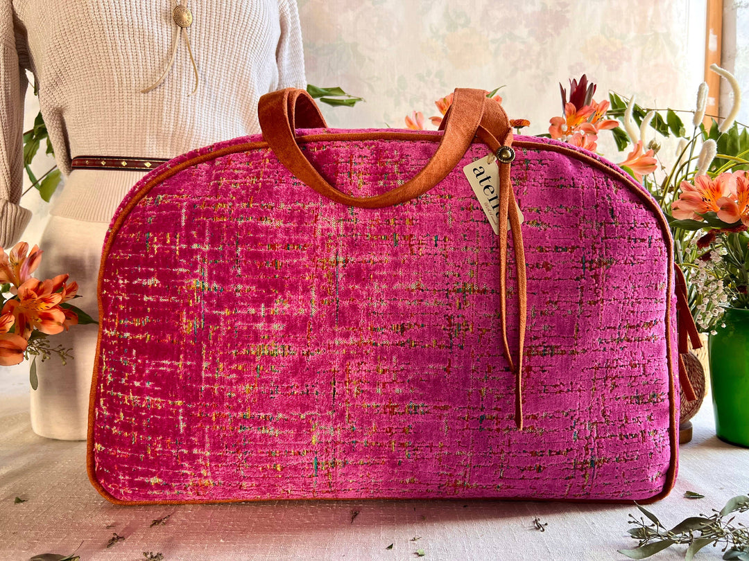A bright pink textured tweed travel bag with brown leather handles and trim, displayed on a table with a cream-colored dress form and flowers in the background.