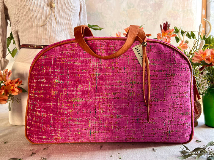 A bright pink textured tweed travel bag with brown leather handles and trim, displayed on a table with a cream-colored dress form and flowers in the background.