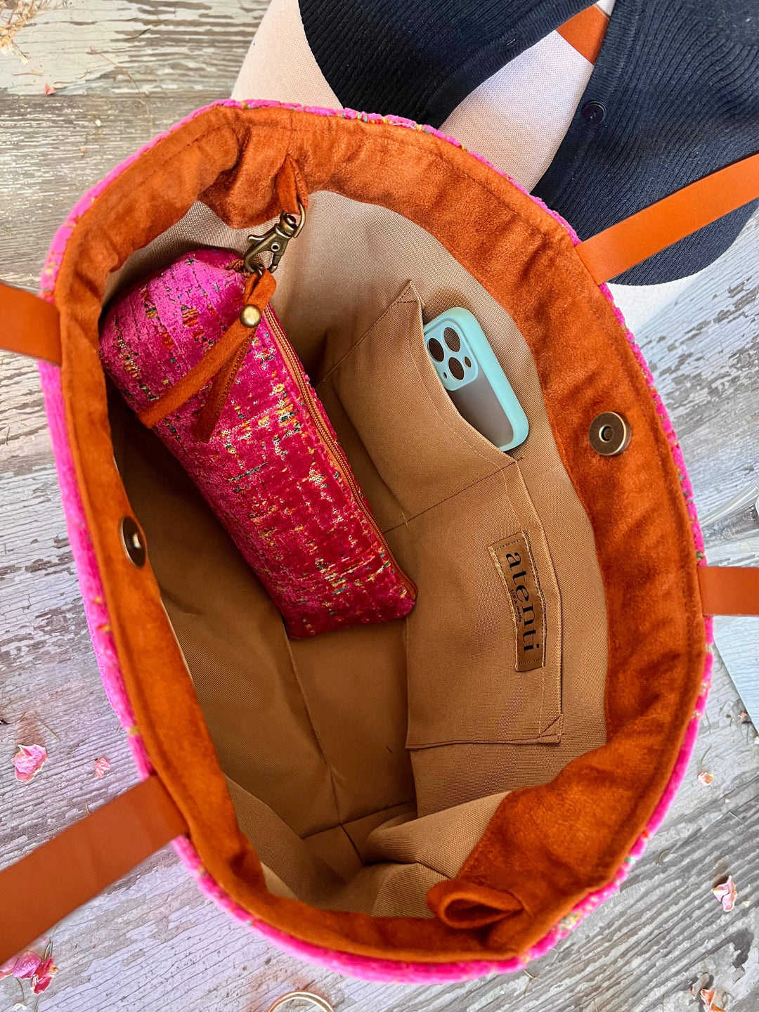 An overhead view of a tan fabric tote bag with a bright pink fuzzy trim and orange-brown handles, shown on a weathered wood surface. A pink patterned zip pouch and a light blue smartphone are placed inside the bag.