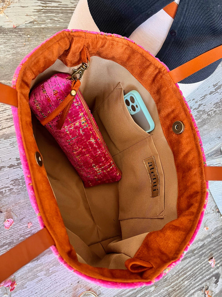 An overhead view of a tan fabric tote bag with a bright pink fuzzy trim and orange-brown handles, shown on a weathered wood surface. A pink patterned zip pouch and a light blue smartphone are placed inside the bag.