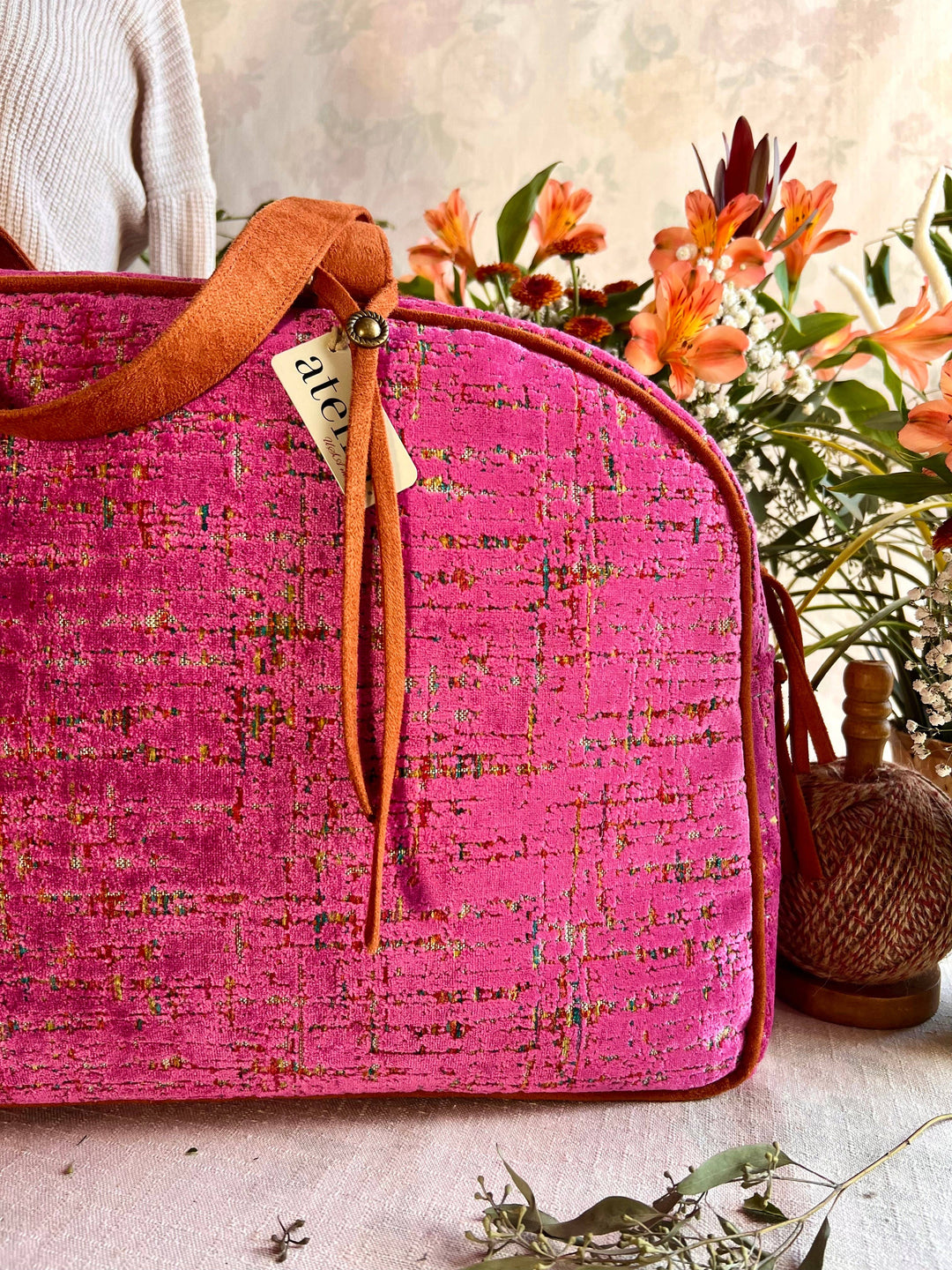 A close-up of a bright pink textured fabric bag with an orange-brown leather strap and a small hanging tag, styled with flowers and dried greenery in a warm indoor setting.