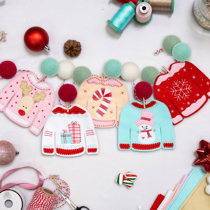 Small embroidered sweaters hanging on colorful ball garland resting on a white surface with Christmas colored notions on the table. 