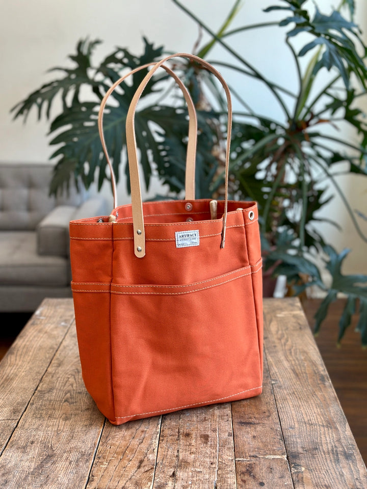 Orange tote bag with leather handles on a wooden table, plant and sofa in the background