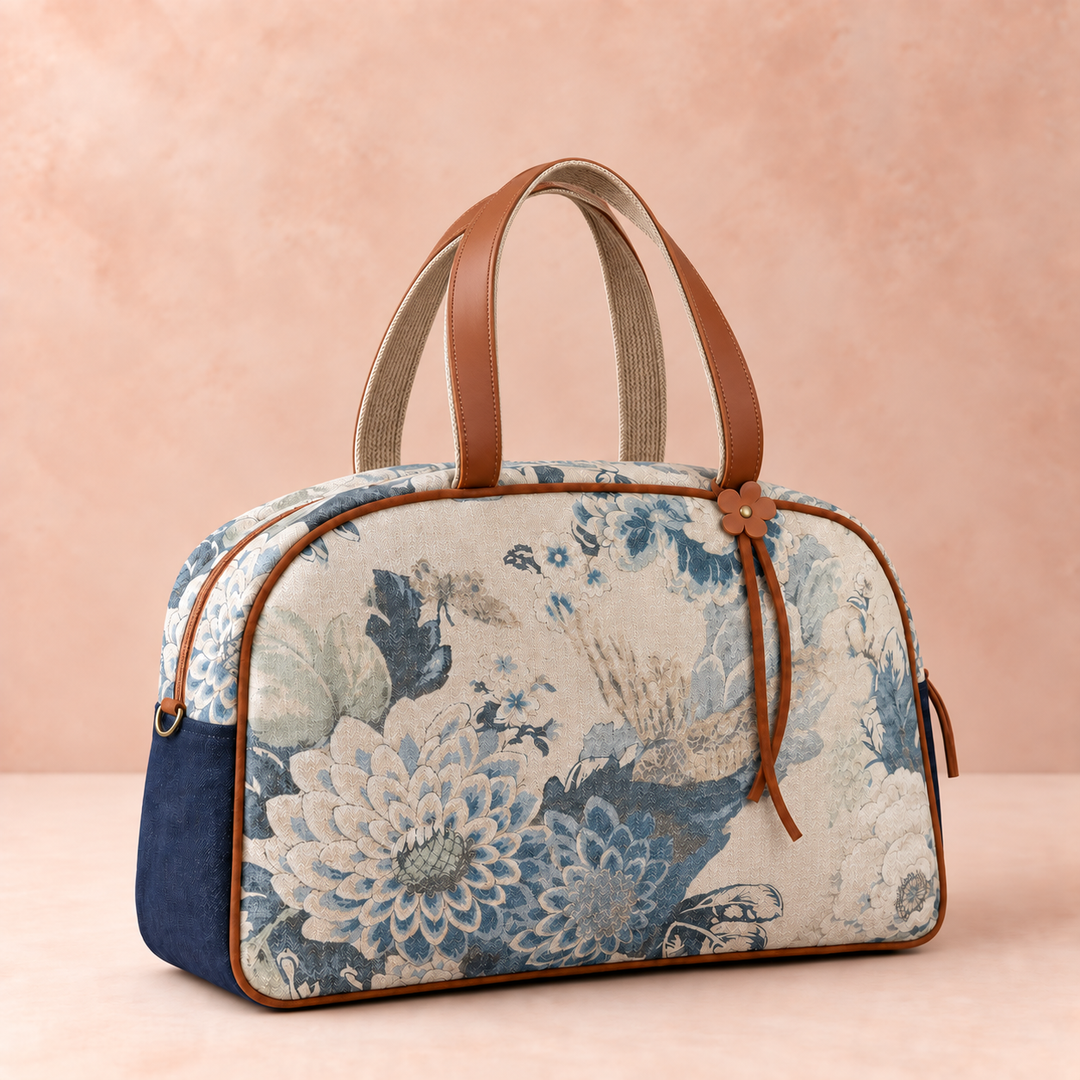 A structured duffel-style handbag with a blue and beige floral jacquard pattern, navy blue side panels, and brown leather handles, set against a soft peach-pink textured background.