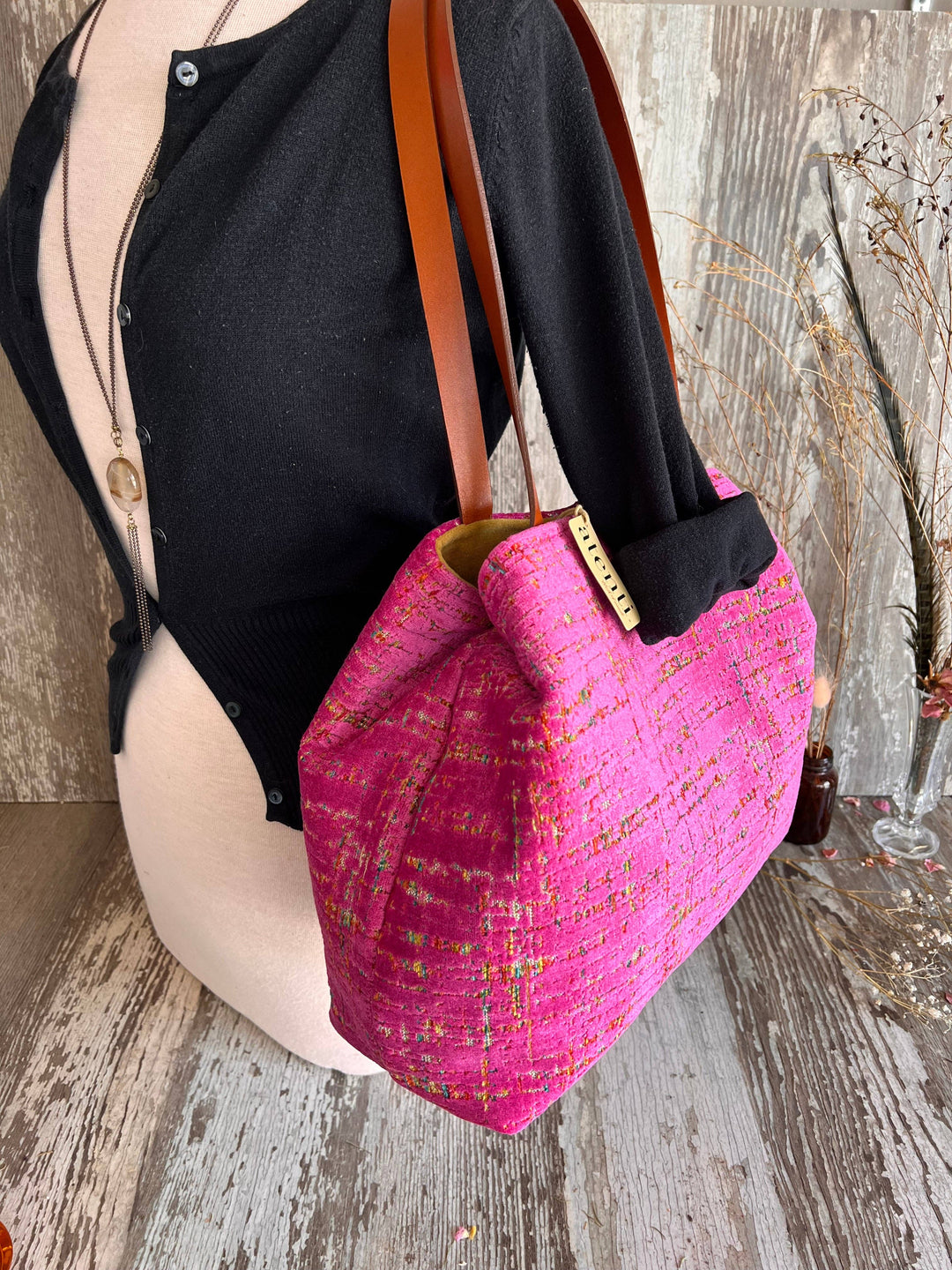 A bright pink woven tote bag with brown leather shoulder straps, displayed on a mannequin torso against a rustic wood background.