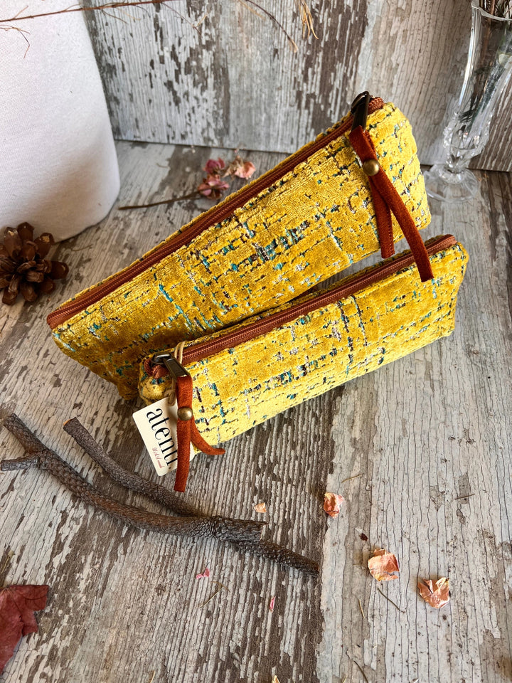 A yellow woven fabric zippered pouch with brown leather-like zipper pulls and a small hanging tag, photographed on a rustic weathered wood surface with dried leaves and branches.