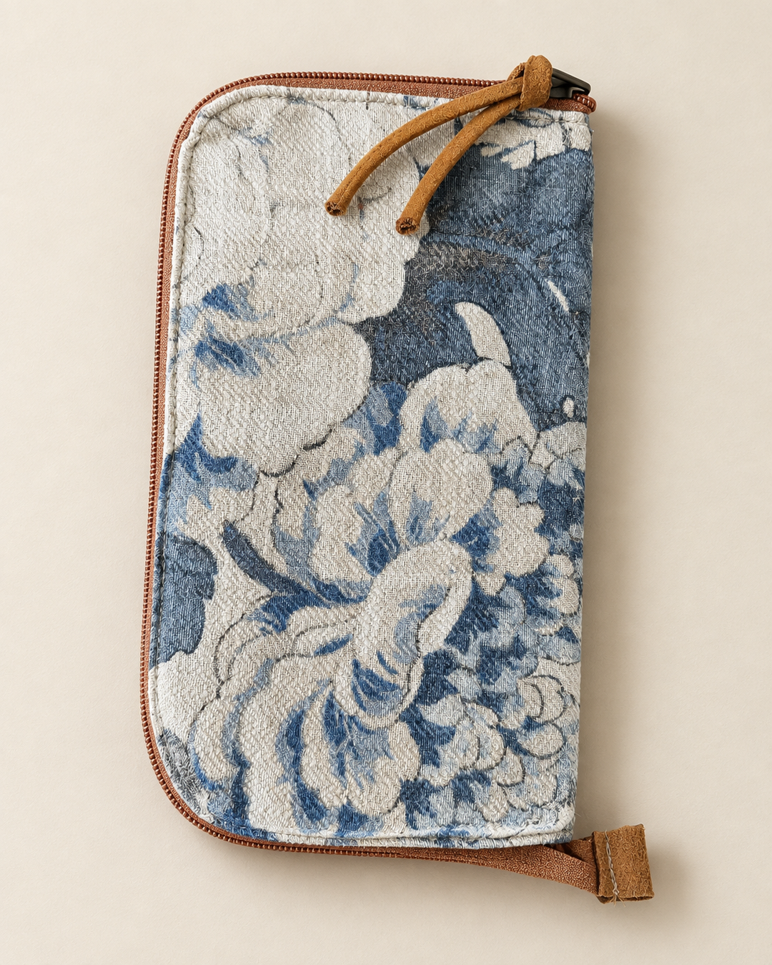 A zip-around clutch wallet featuring a blue and white floral tapestry pattern, brown leather trim, and a matching leather wristlet strap, shown on a neutral beige background.