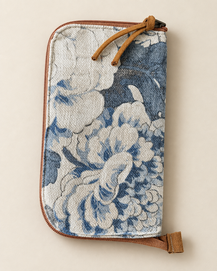 A zip-around clutch wallet featuring a blue and white floral tapestry pattern, brown leather trim, and a matching leather wristlet strap, shown on a neutral beige background.