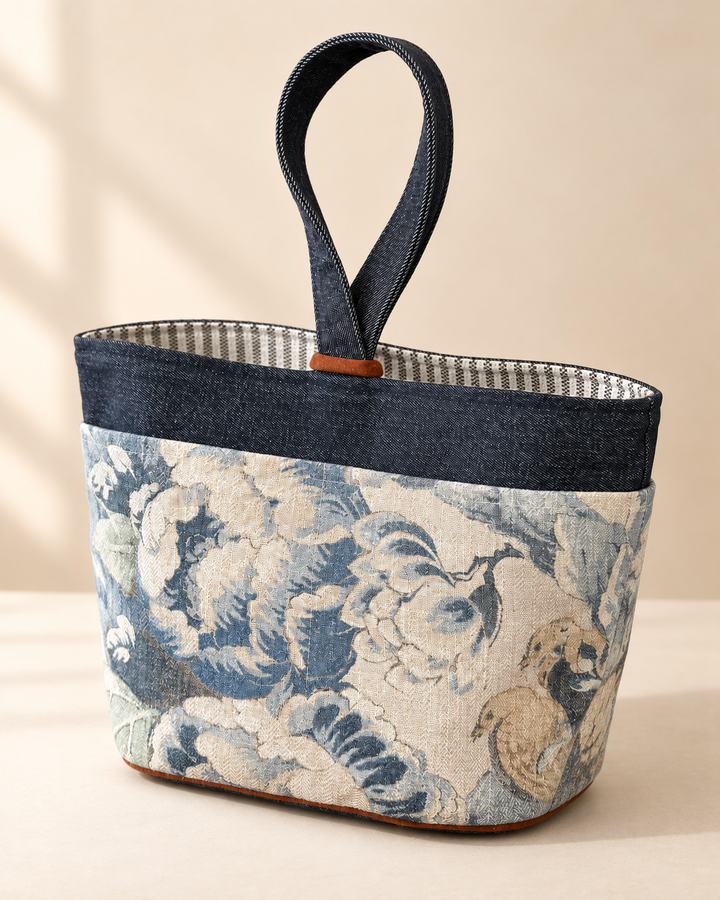 A structured tote bag with a dark navy blue top band and handle, featuring a blue and beige floral toile-style print on the lower body, set against a warm beige background.