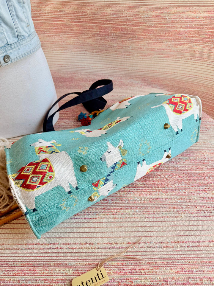 Bottom of a blue tote bag with llamas on it, there are four gold feet  on the bottom of the bag. 