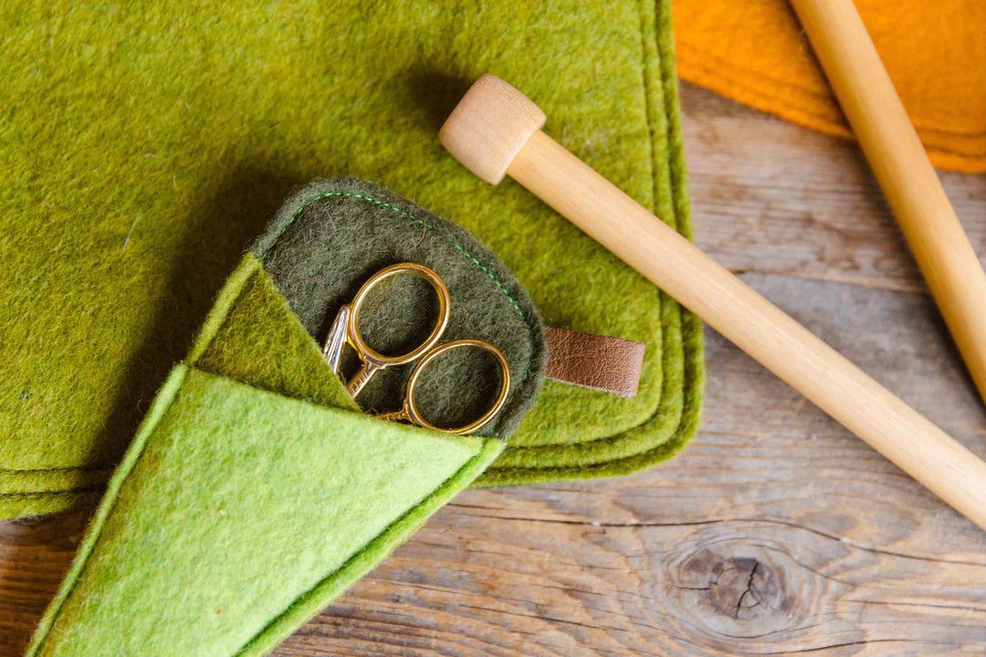 A close-up of a green felt sewing or craft pouch holding a pair of gold scissors, with wooden knitting needles resting on a rustic wood surface.
