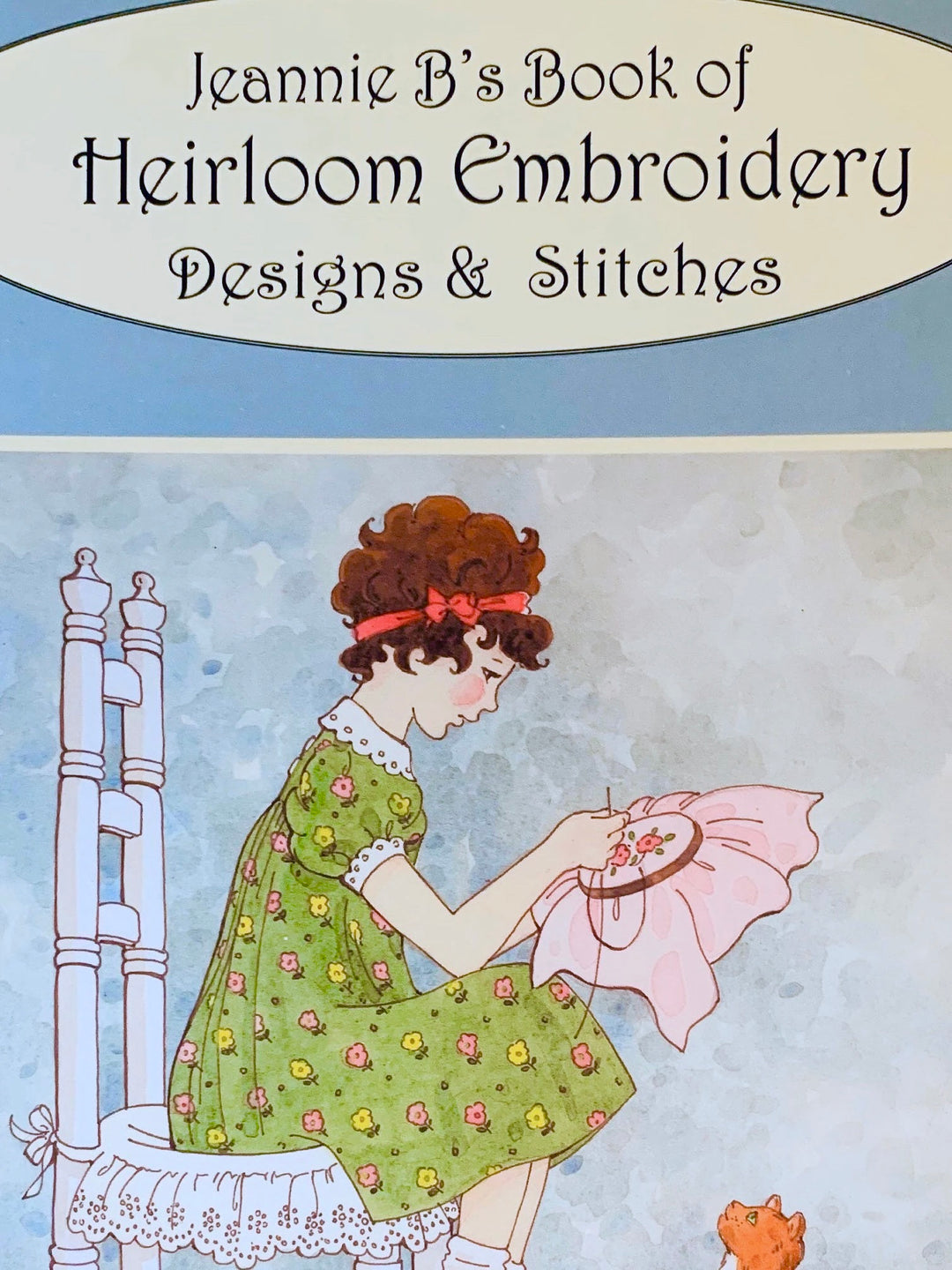Cover of a book titled 'Jeannie B's Book of Heirloom Embroidery Designs & Stitches' featuring an illustration of a girl sitting at a table with embroidery hoop.