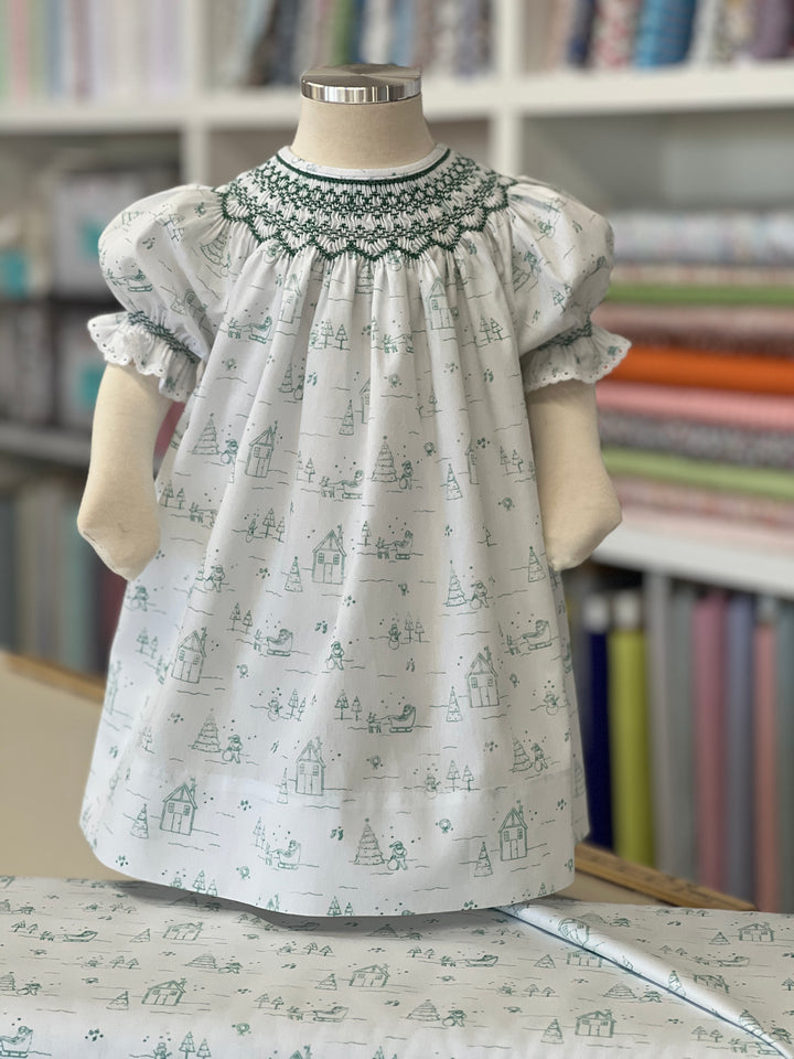 Bishop dress smocked in forrest green and the fabric has Christmas scenes printed on a white background.