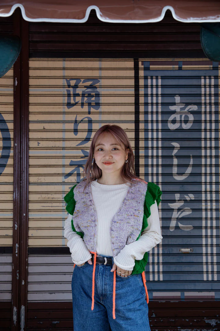 A young Japanese woman stand in front of a store front, and she's wearing a heathered purple and green ruffled knitted sweater with orange drawstrings.