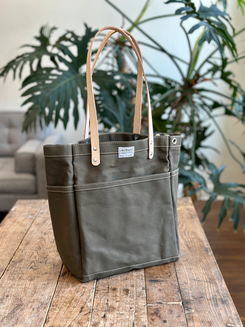 Green tote bag with brown leather straps on a wooden surface with plants in the background