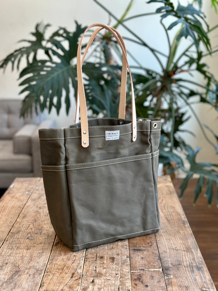 Green tote bag with brown leather straps on a wooden surface with plants in the background